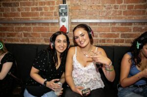 Two friends smiling in silent disco headphones by a brick wall