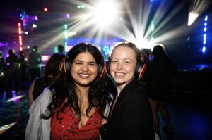 Two friends smiling in headphones in front of stage light beams