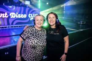 Two friends in headphones in front of the Silent Disco Club sign